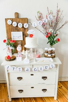 a table topped with cake and cupcakes on top of a white dresser next to flowers