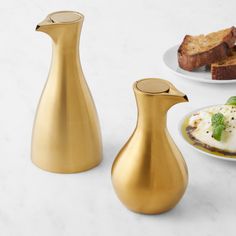 three gold vases sitting next to each other on a white counter top with plates and bread