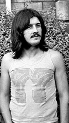 a man with long hair and mustache standing in front of a bush wearing a tank top