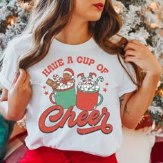 Our cup of cheer Christmas shirt makes the perfect addition to your Holiday collection! This shirt is a unisex fit so please check the sizing chart in the photos section for exact measurements.  How we make our shirts - The design shown is printed directly onto the shirt using a professional grade DTG printing machine that allows the ink to absorb into the fabric. This allows your shirt to last longer and will prevent cracking or peeling over time. You will love it! If you have any questions ple Cute Christmas T-shirts, Trendy Christmas Shirts, White Pre-shrunk Christmas Tops, Christmas Graphic Tee, Christmas Tshirt Designs, Christmas Shirt Women, Holiday Tees, Hot Cocoa Christmas, Cup Of Cheer