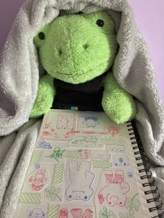 a green stuffed animal sitting under a blanket on top of a notebook with stickers