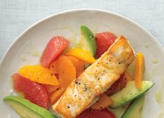 a white plate topped with fish covered in citrus and avocado wedges next to sliced oranges