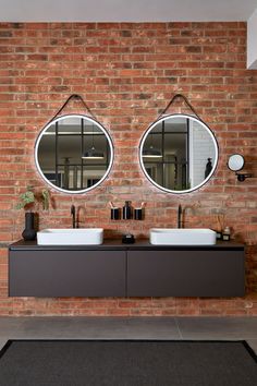 two round mirrors hanging on the side of a brick wall next to double sinks in a bathroom