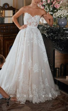 a woman standing next to a fireplace wearing a wedding dress with white flowers on it