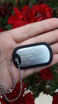 a hand holding a black and silver dog tag on it's side with red flowers in the background