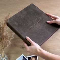 a person holding a brown book on top of a wooden table next to two photos