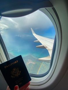 a person holding up a passport and looking out an airplane window at the ocean below