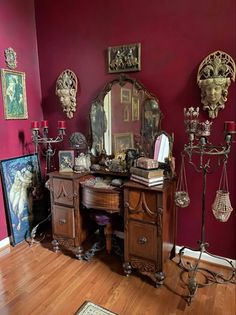 an antique desk and mirror in the corner of a room with red walls, wood floors and artwork on the wall