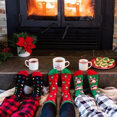 three people sitting in front of a fireplace with christmas socks and mugs on their feet