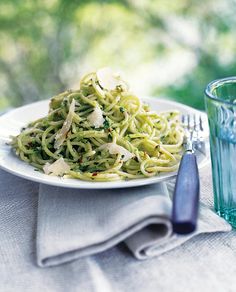 a white plate topped with pasta covered in pesto and parmesan cheese next to a glass of water