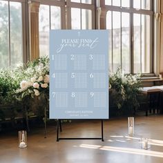 a blue wedding seating sign with white flowers and greenery in front of large windows