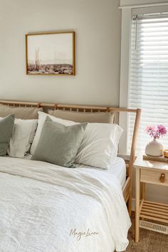 a bed with white sheets and pillows next to a wooden night stand in front of a window