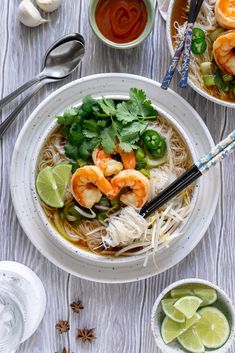 two bowls filled with shrimp and noodles on top of a white wooden table next to spoons