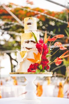 a three tiered cake with flowers on top is sitting on a white table in front of an orange and red flower arrangement