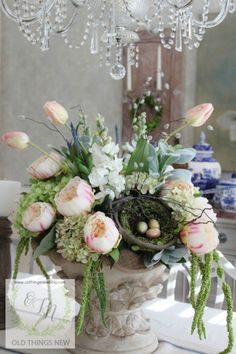 a vase filled with flowers and greenery on top of a white table next to a chandelier