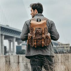 a man with a brown backpack is walking in tall grass near a bridge and water