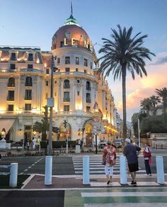 people are standing on the sidewalk in front of a large white building with palm trees