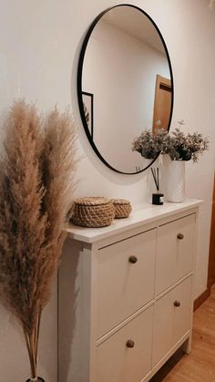 a white dresser topped with a round mirror next to a vase filled with dried plants