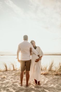 a man and woman standing on top of a sandy beach next to the ocean at sunset