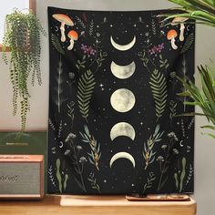 a tapestry with three phases of the moon and plants in front of it on a shelf