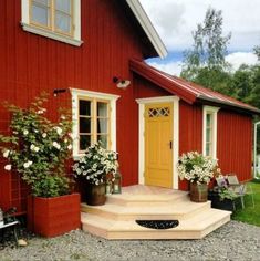 a red house with white flowers and potted plants on the front porch, next to a yellow door