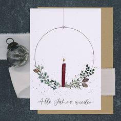 a christmas card with a candle and pinecone wreath on it, next to a glass ornament