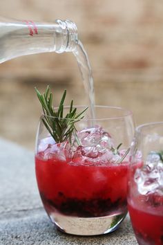 a glass filled with red liquid being poured into another glass and garnished with rosemary