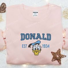 Introducing the Donald Est 1934 Embroidered Shirt, a timeless piece that pays homage to the iconic Disney character. Made with premium quality fabric, this shirt showcases intricate embroidery of Donald Duck, adding a touch of nostalgia to your wardrobe. With its classic design and comfortable fit, it is perfect for casual outings or Disney-themed events. [...] Unique Clothing Stores, Gifts For Disney Lovers, Embroidered Apparel, Maroon Hoodie, Duck Cloth, Best Gift Ideas, Disney Character, Unique Clothing, Hoodie Material