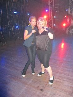 two women standing on a wooden floor in front of a stage with lights and microphones