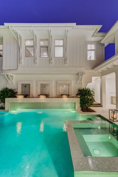 an empty swimming pool in front of a large white house at night with lights on