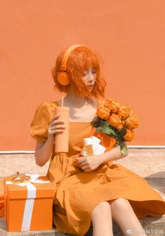 a woman with red hair sitting on the ground next to a gift box and orange flowers