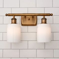 two lights are mounted on the wall next to white tiled walls in a bathroom with gold fixtures