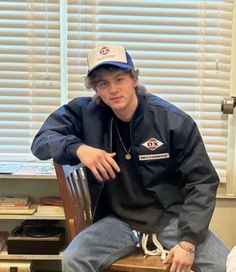 a young man sitting on top of a wooden chair wearing a blue jacket and hat