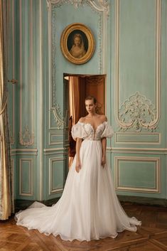 a woman in a white wedding dress standing in front of a blue wall with ornate paintings on it