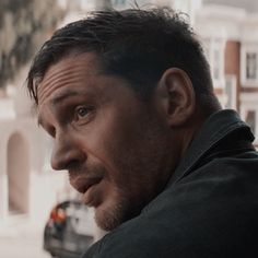 a man looking off into the distance with buildings in the background and cars parked on the street behind him