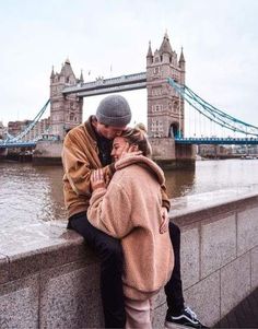 a man and woman sitting next to each other on a wall near the water with a bridge in the background