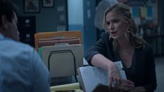 a woman sitting at a desk talking to a man who is holding papers in his hand