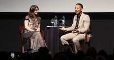 two people sitting on chairs in front of a screen talking to each other at an event