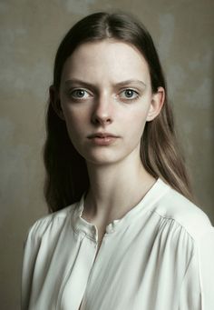 a woman with long hair wearing a white shirt and looking at the camera while standing in front of a wall