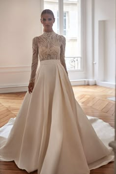 a woman in a long sleeved wedding dress stands on a wooden floor with her hands behind her back