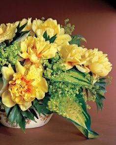 a white vase filled with yellow flowers on top of a brown table next to a green plant