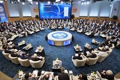 a large group of people sitting at tables in front of a big screen with a blue carpet