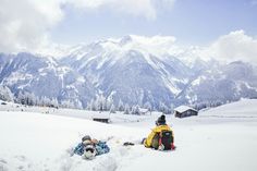 two people sitting in the snow on top of a snowy hill with mountains in the background