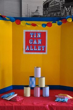 a red table topped with rolls of toilet paper next to a yellow wall and a sign that says tin can alley