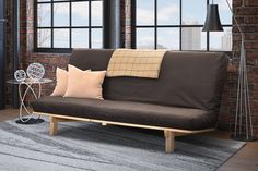 a brown futon couch sitting in front of a window next to a table with two pillows