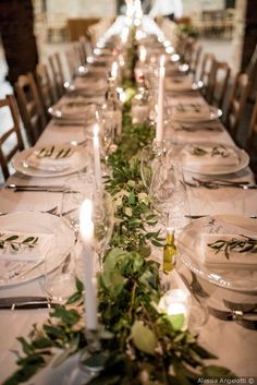 a long table is set with white plates and silverware, greenery and candles