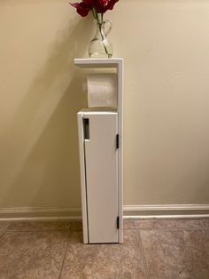 a vase with flowers on top of a white cabinet next to a wall mounted toilet paper dispenser