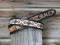 a pair of black and white leather belts with an embroidered design on the bottom, sitting against a wooden fence post