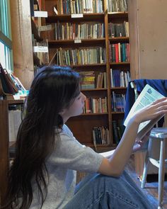 a woman sitting in front of a bookshelf holding a book and looking at it