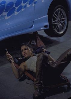 a woman laying on the ground next to a blue car holding a cell phone in her hand